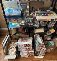 Wide view of two shelves with various art supplies, boxes of paints, brushes, art books, and a large box of sea shells on floor.