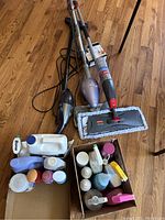 Photo showing the steam mop, handheld devices, mop pads, and two boxes with various cleaning bottles and containers on a hardwood floor.