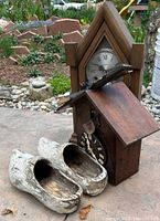 Photo showing vintage wooden cuckoo clock, wooden mantle clock, and pair of rustic wooden shoes on stone and concrete outdoor surface, with garden background.