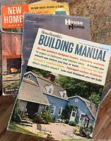Vintage home design magazines on a rough surface, featuring House Beautiful's Building Manual with a blue house on the cover.