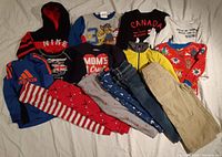 Full view of assorted boys clothing items laid out on a white surface showing hoodies, tops, and pants in various colors and prints.