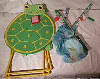 View of Sunny Patch Frog Bean Bag Toss target board showing frog design with numbered sections, some damage to yellow metal frame, one red bean bag included, and Catch and Count Fishing Game rods and mesh net visible