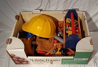 Box of assorted toy tools including yellow toy hard hat, various handheld tools, and fabric tool belt packed in a white box.