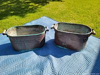 Two antique copper boilers placed on a blue protective cloth outdoors on grass, showing overall size and shape.