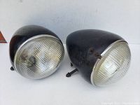 Photo of two black torpedo-style antique car headlights showing front textured glass lenses and metal mounting rods