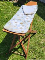 Full view of the antique wooden ironing board standing on grass, showing padded floral cover and folded wooden legs.