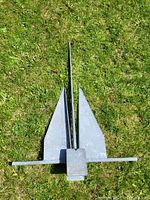 Top view of large metal boat anchor laid flat on grass, showing the two flukes, shank and crossbar.