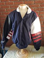 Front and left side view of the Goodwrench promo jacket on a mannequin against a brick wall, showing navy blue body with white and red accented sleeves and white collar.