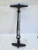 Front view of Raleigh air pump showing metal cylindrical body with black 'Raleigh' branding, black T-handle, footrests, and pressure gauge at base.