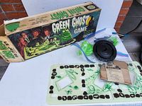 Photo showing the game box, foldable game board, multiple green ghost masks still attached to plastic frames, black circular device, and instruction manual.