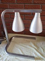 Side angled view of dual conical silver metal heat lamps mounted on an L-shaped metal heating light frame against a red brick wall background.