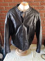 Front view of vintage black leather motorcycle jacket on mannequin against brick wall. Shows buttoned collar, zipper, zippered pockets, and surfaces of jacket.