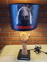 Photo showing full lamp with zombie hand base and graphic shade with Walking Dead logo, lit and unlit