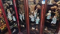 Front view of four-panel black lacquered room divider with multicolored 3D figures and gold-painted landscape scenes