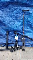 Three black guitar stands laid on blue tarp and ground surface.
