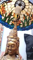 Close up of Quan-Yin statue's head and upper torso under Tiffany style stained glass shade showing floral motifs and light bulb.
