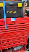 Black Stanley portable toolbox with yellow latches and handle, strapped to the top of a red metal rolling toolbox.