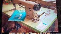 Top-down angled view showing Juki DDL-552 sewing machine on working surface of wood and metal table, with blue Juki instruction manual beside machine, oil cans in background.