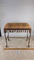 Front view of stool showing upholstered seat with patterned fabric and tassel fringe, wrought-iron curved legs.
