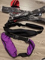 Money belts arranged on a wooden floor, some purple with adjustable black straps and plastic buckles, others sealed in plastic wrapping.