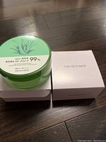 Two jars of The Face Shop Jeju Aloe 99% soothing gel in green and white packaging displayed with respective white boxes on dark wooden surface.