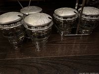 Two sets of vintage glass condiment servers on a dark wooden surface. First set with three containers in a metal holder, second set with two containers in a metal rack.