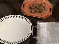 All three trays shown together: orange painted floral tray at top, white cut-out oval tray bottom left, silver embossed tray bottom right.