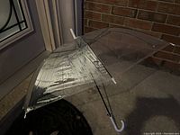 Clear transparent bubble umbrella open showing dome-shaped canopy and white hooked handle on a porch.