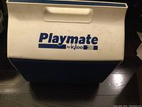 Exterior view of the Igloo Playmate cooler showing the lid with the Playmate by Igloo logo in blue on white plastic. Cooler has blue body portion visible along the edges.
