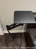 Adjustable black laptop stand mounted on a black arm with hinges, positioned beside a dark sofa, showing the platform surface and arm’s bending structure.