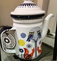 White enamel coffee pot with colorful folklore design, showing the handle with tag and spout.
