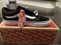 Profile side view of black and white Vans Old Skool sneaker on top of Vans branded box, tag attached to shoe.
