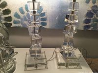 Pair of clear crystal lamp bases featuring stacked cubic shapes with some visible small chips on base and column, photographed from front.