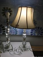 Pair of crystal lamps shown side by side on surface against floral wallpaper background. One lamp includes beige fabric shade with trim, the other is lamp base only.