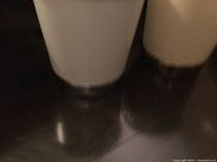 Close-up photo showing the white metal trash bin with chrome bottom on the left and beige bin with gold bottom on the right, placed on a reflective surface.