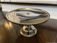 Photo of shiny stainless steel cake stand on wooden floor, showing full top and pedestal base.
