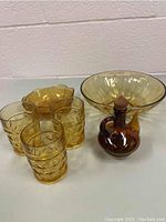 Full set of amber glass pieces on a white surface against a wall: four textured tumblers, small scalloped bowl, large ribbed bowl, and amber bottle with wooden stopper.
