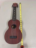 Soprano ukulele positioned next to a measuring tape to indicate size, showing wooden body and strings.