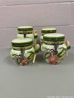 Four ceramic canisters displayed with green lids, secured with metal clasps, featuring painted fruit designs and attached spoons