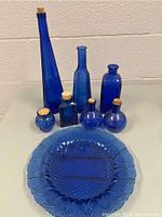 Front view of 7 cobalt blue glass bottles with cork stoppers and 1 cobalt blue glass plate on table.