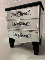 Front and side view of mirrored jewelry box with three drawers, each with decorative black floral pattern and clear crystal-like knobs, standing on black legs