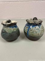 Pair of small pottery dishes with bulbous shapes and lidded tops showing glazed finishes in blue, green and beige colors with rustic metal-like lids.