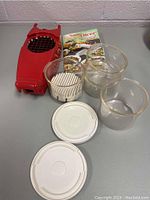 Overview of the vintage Genius Nicer Dicer set including the red dicing unit, clear containers, white lids, and instruction booklet laid out on table.