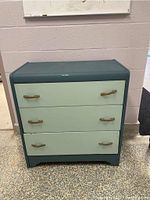 Full view of a wood dresser with three drawers, showing the painted teal body and pale green drawer fronts with wooden handles