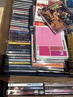 Box full of music CDs including artists and various albums, showing CD jewel cases stacked together.
