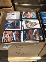 Top-down photo of a box filled with various DVD and CD cases, showing the lot contents and condition.