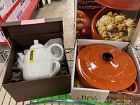 Orange enameled cast iron hot dip baker in open box, showing lid and side handles, packaged with seasoning inside.