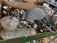 Box with assorted kitchenware items including strainers, funnel, metal containers, glass dishes shown piled inside a cardboard box