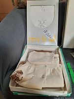 Cream-colored double-breasted knit cardigan with embroidered girl motif and buttons, partially folded inside an open box with brand name 'mab'