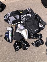 Photo showing the overall hockey gear lot including pants, elbow pads, gloves, and the hockey bag on asphalt.